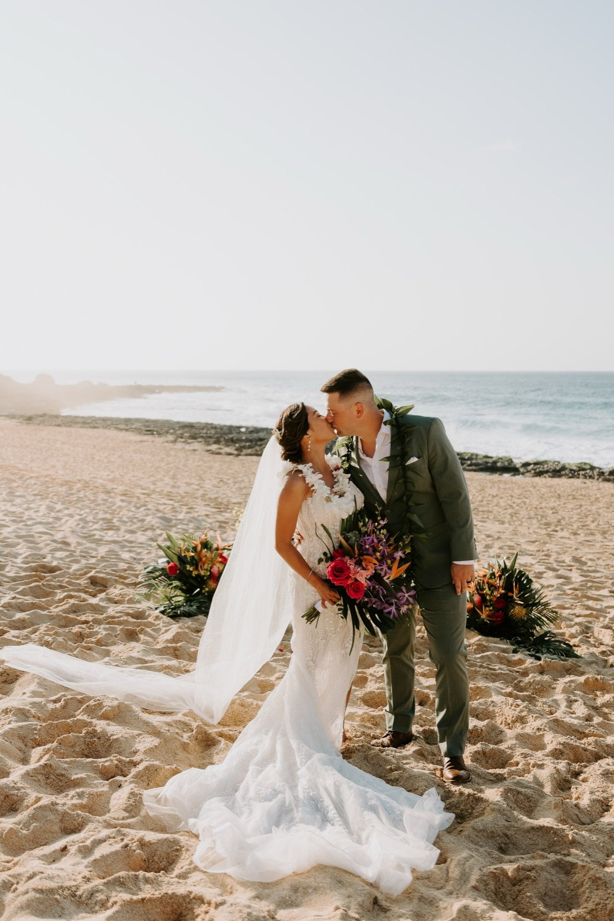 A kiss on the sand at golden hour, basket arrangements marking the aisle behind.