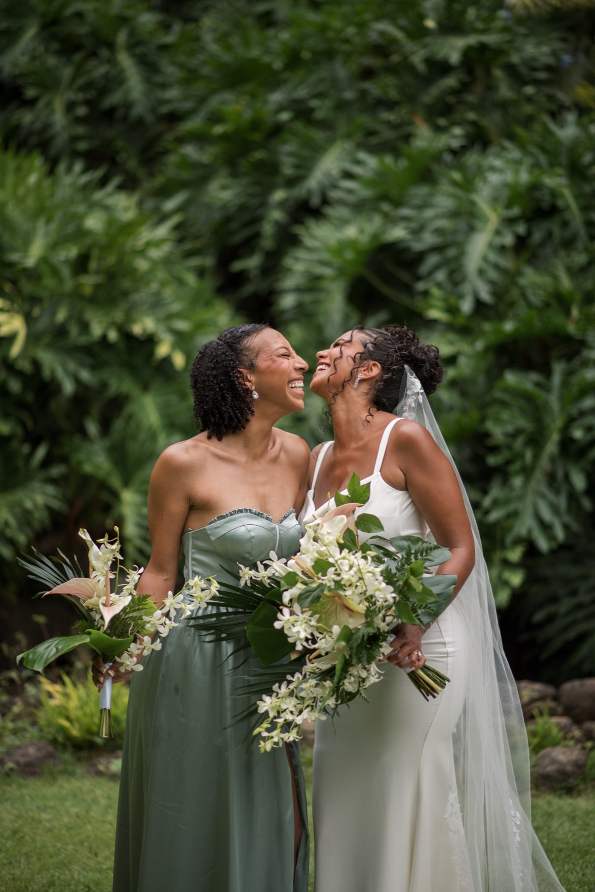A bride and her bridesmaid laughing, a bouquet of white orchid, anthurium, and monstera between them.
